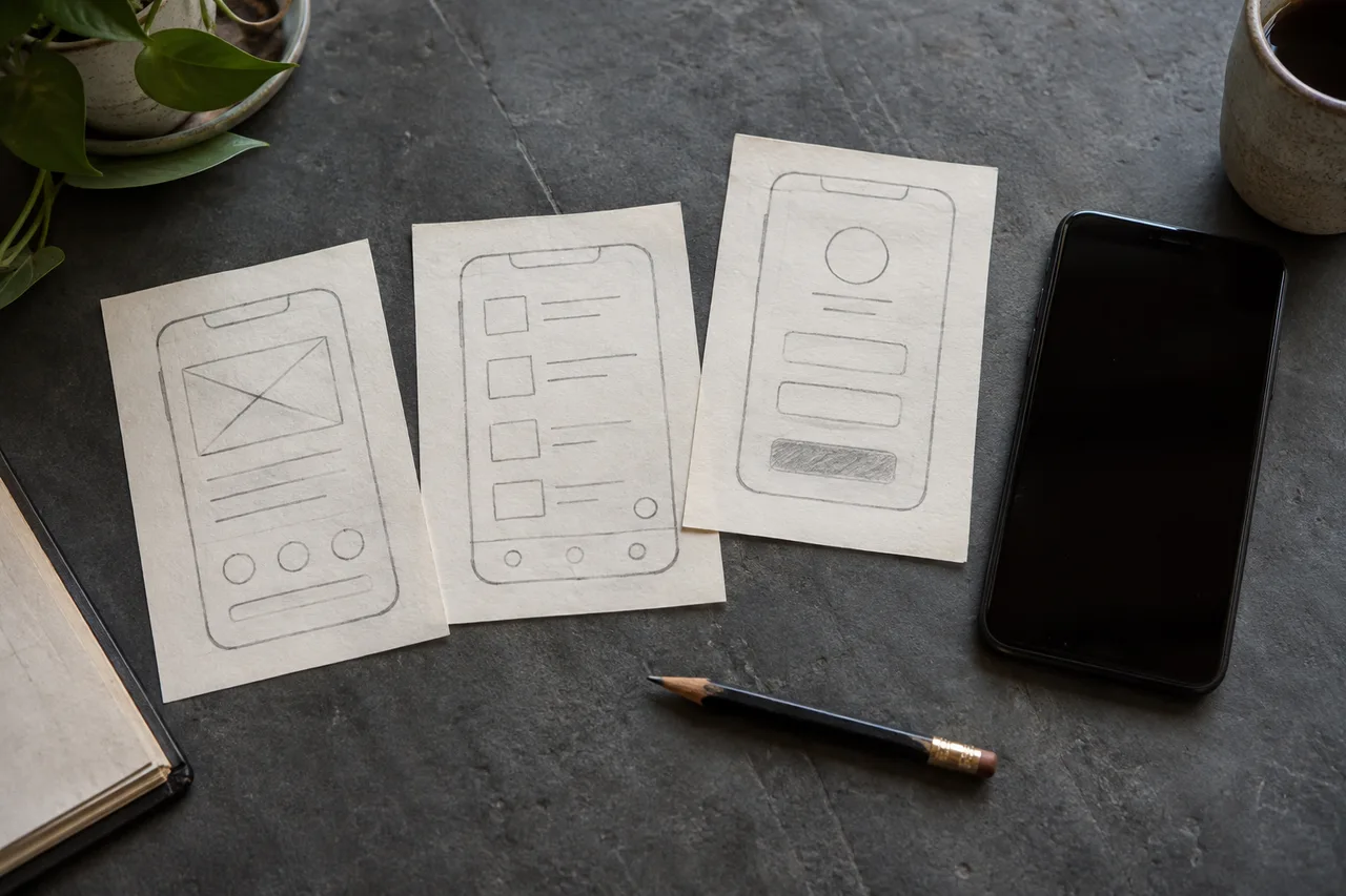 Three paper phone-screen prototypes beside a black smartphone and pencil.